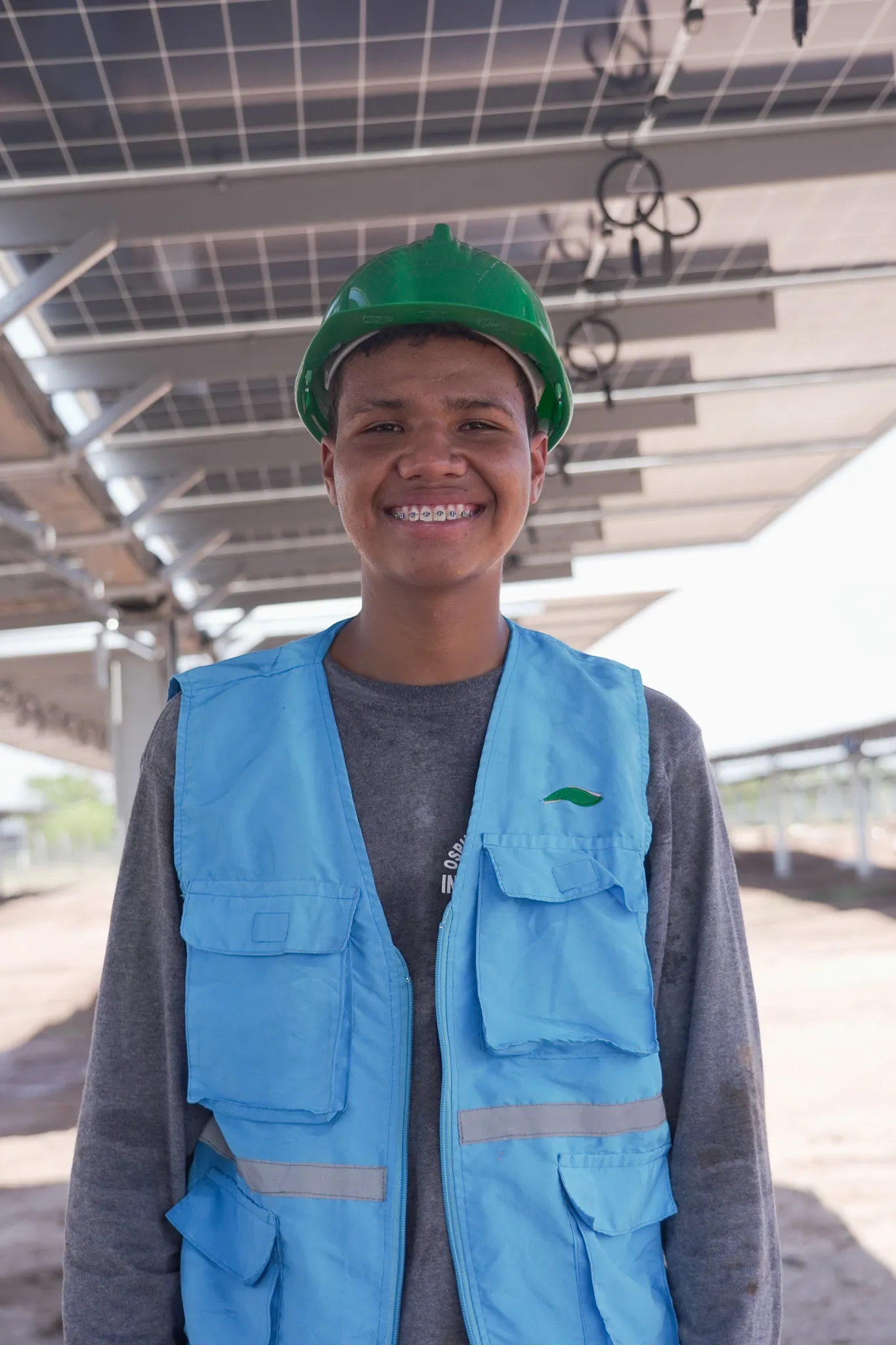 Retrato de un joven sonriente con chaleco azul de seguridad y casco verde, posando frente a una instalación de paneles solares.